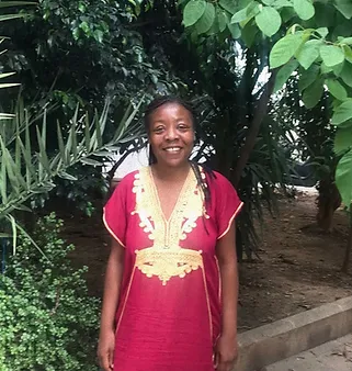 A woman in a red dress with gold embroidery smiles in a lush garden surrounded by green plants. This image truly embodies the essence of "About Me."