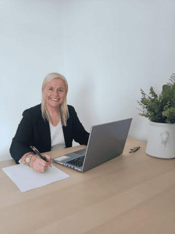 Una mujer profesional sonríe a la cámara mientras está sentada en un escritorio con una computadora portátil, papel, bolígrafo y plantas decorativas en una oficina luminosa, lista para colaboraciones.