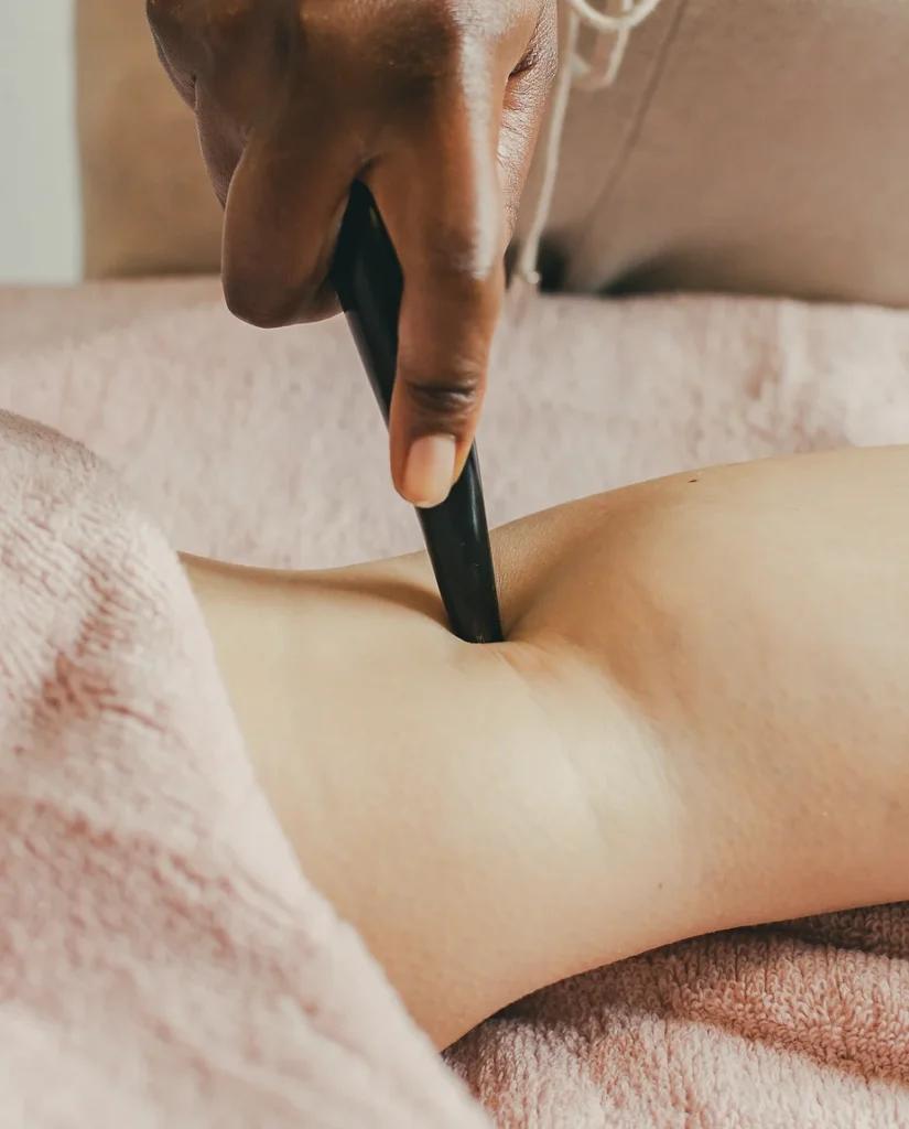 A therapist uses a black massage tool on a client's bare back, focusing on a specific point in the deep tissue. The client is lying down, covered with a pink towel.