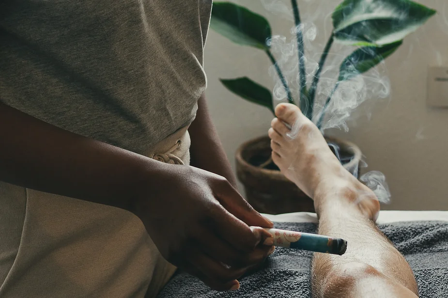 A person sitting cross-legged, using moxibustion, a traditional therapy that involves a smoking stick held close to the knee, emitting smoke, in a serene and relaxing environment with a plant in the background.