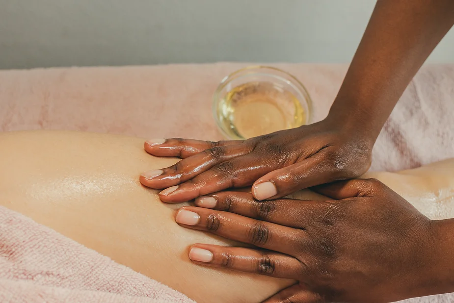 Hands applying anti-cellulite oil to a person's back during a massage treatment, with a container of visible oil next to them.