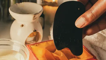 A close-up of a hand holding a small black gua sha tool on a warmly lit table with a ceramic bowl and cloth. The scene conveys a soothing atmosphere, probably related to skin care or