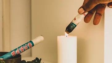 A person lights a white candle with a decorated fireworks stick, placed next to other similar fireworks and a bowl, on a beige background.