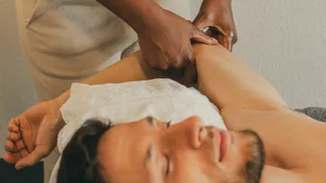 A masseuse massages the arm of a relaxed man lying on a massage table, who looks happy and smiling. The surroundings look relaxing and professional, perfect for a Holistic Rel Massage.