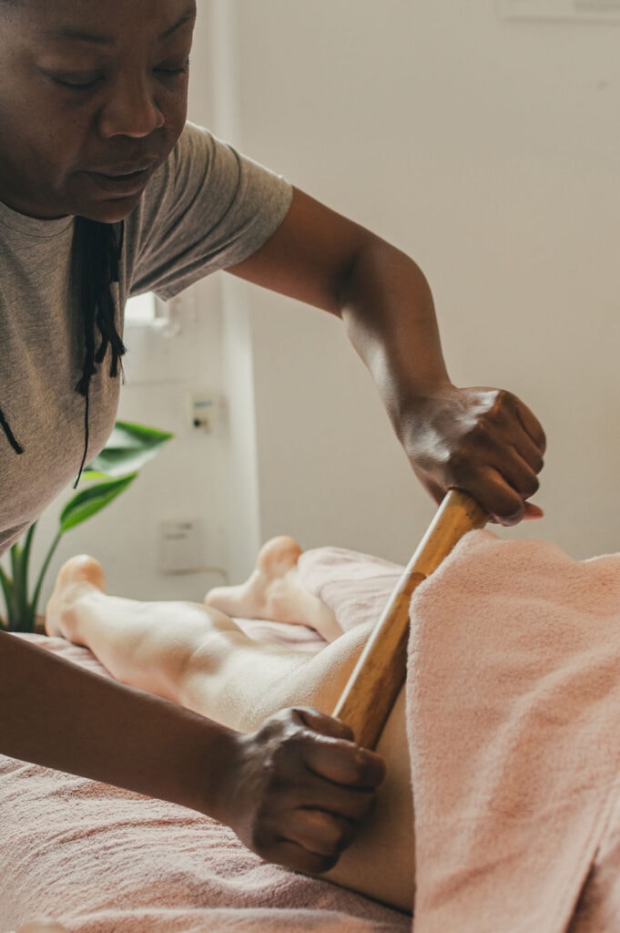 A woman performs an anti-cellulite massage using a wooden tool on the back of a client who is lying face down and covered with a pink towel in a serene environment.