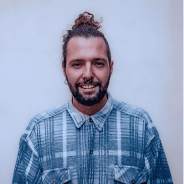 Un hombre sonriente con un moño, barba y una camisa a cuadros de pie sobre un fondo azul claro para colaboraciones artísticas.