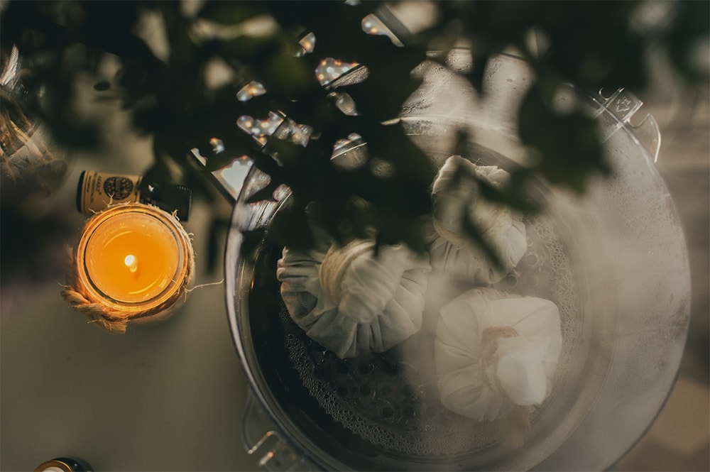 Una imagen serena que presenta una vista en primer plano de una vela brillando junto a flores blancas bajo la sombra de hojas verdes, vista a través de un enfoque suave. La escena crea una atmósfera tranquila e íntima, que recuerda a una