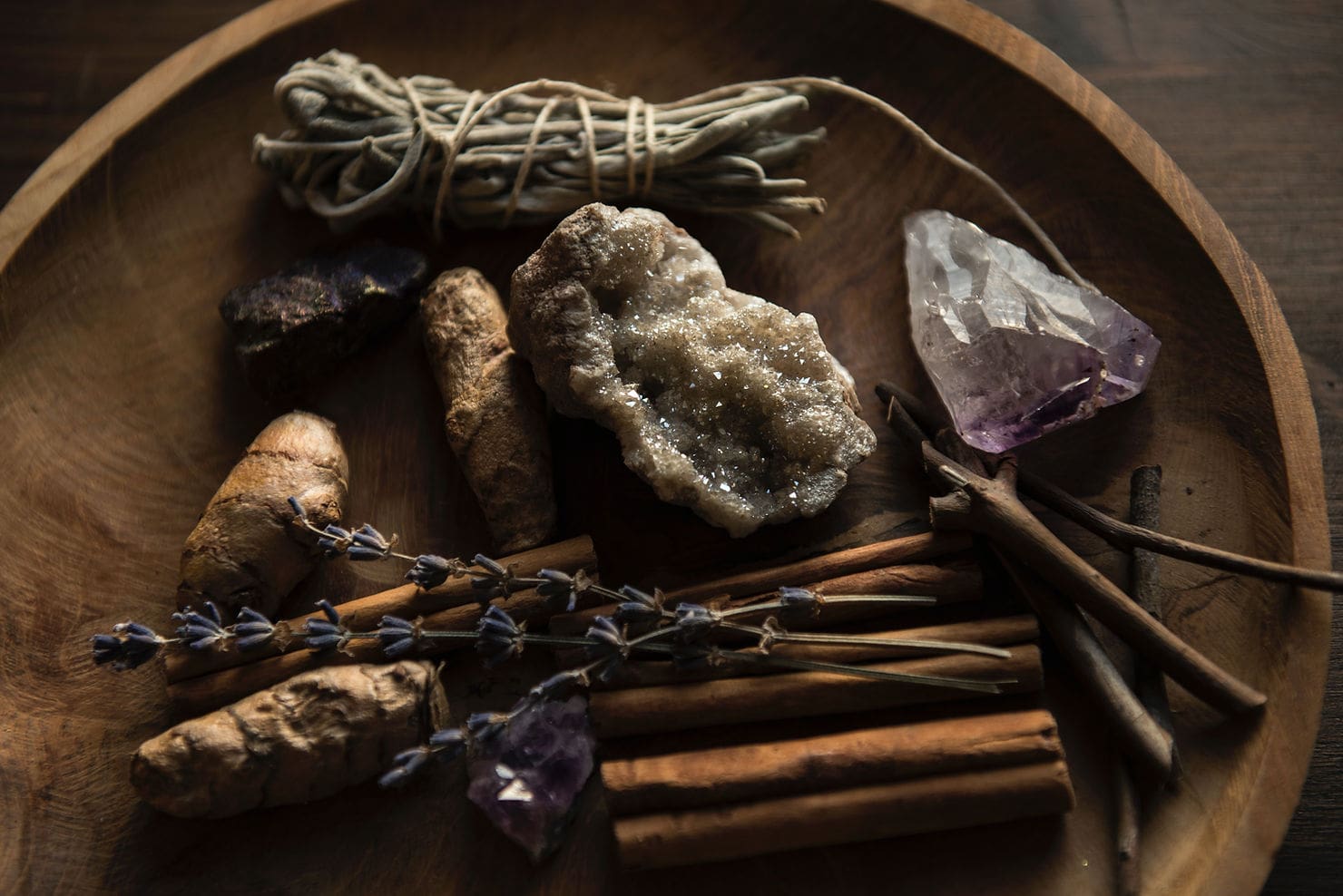 Diversos elementos naturales sobre un plato de madera, como canela en rama, lavanda, cristales, trozos de jengibre y un manojo de cordel dispuestos en un ambiente rústico ideal para técnicas de relajación.