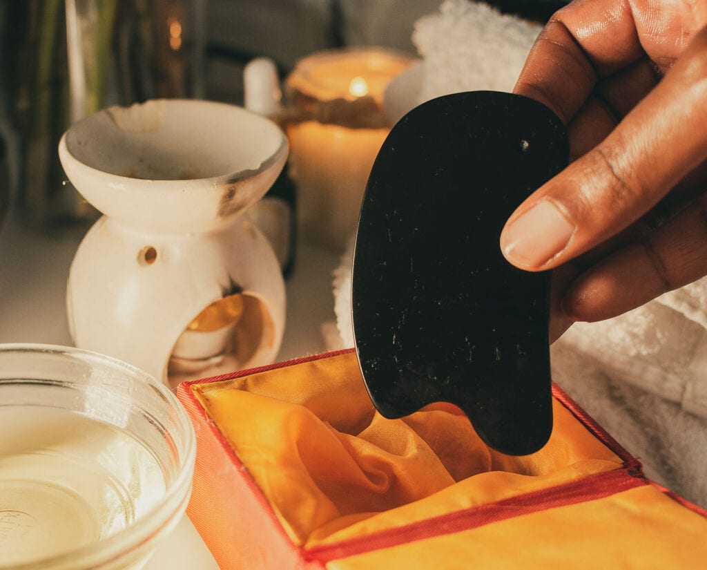 Un primer plano de una mano que sostiene una herramienta Gua Sha negra sobre una bolsa de seda naranja, con un candelabro y elementos de spa al fondo.