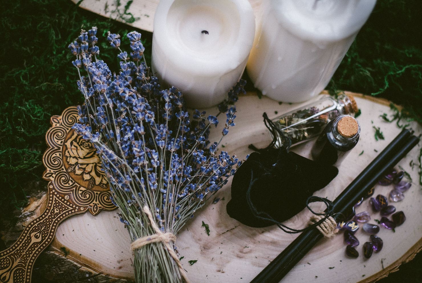 Un arreglo rústico de naturaleza muerta con un ramo de lavanda seca, dos velas blancas, una tabla de madera ornamentada y un pequeño frasco de aceites esenciales con una tapa negra, todo ello contra un musgo.