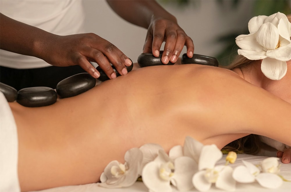 A woman gets a hot stone massage on her back in a serene spa setting, surrounded by white orchids, with a therapist's hands laying smooth stones during her spa treatment.