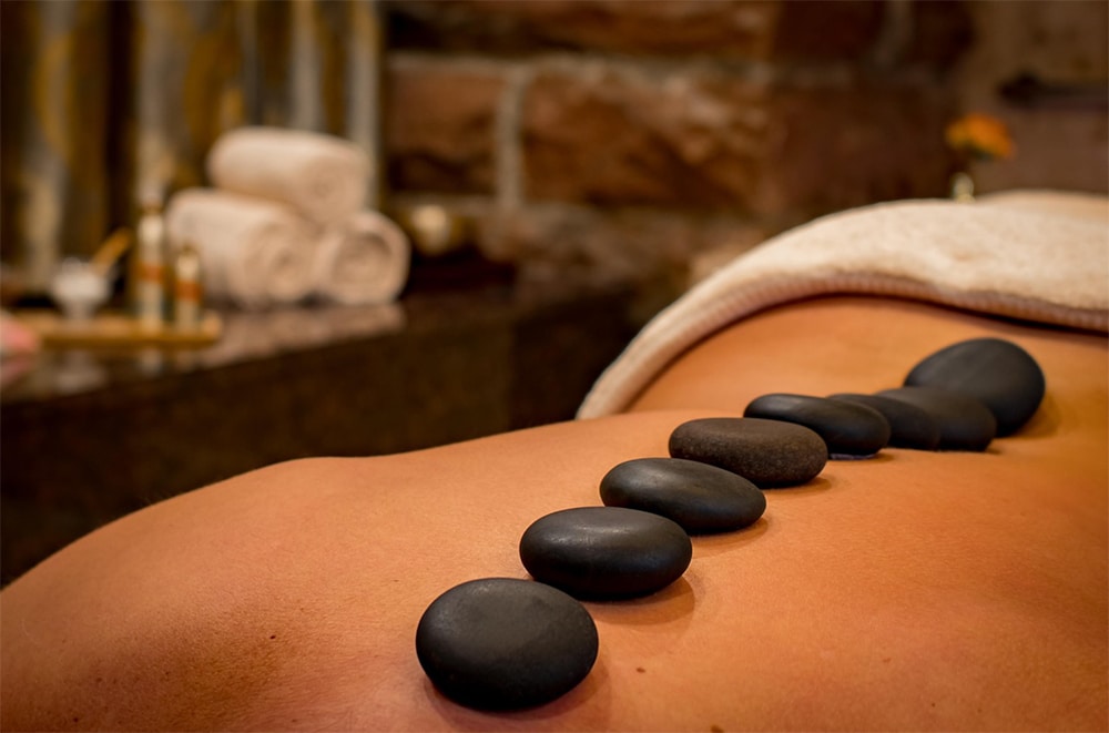 Close-up of a person receiving a hot stone massage, showing soft black stones lined up along their bare back in a serene spa setting.