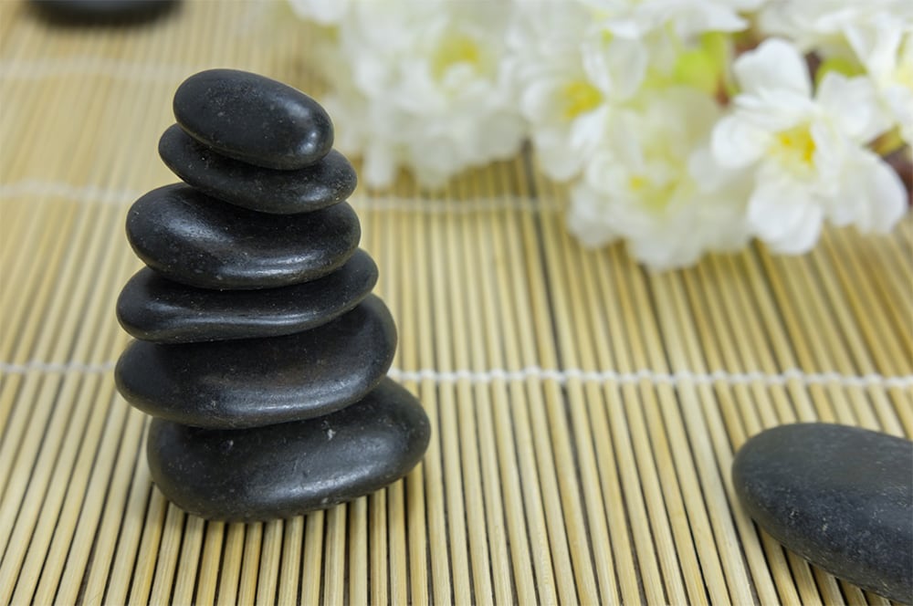 A pile of smooth black stones in a balanced formation on a bamboo mat, ideal for Hot Stone Massage, with blurred white flowers in the background.