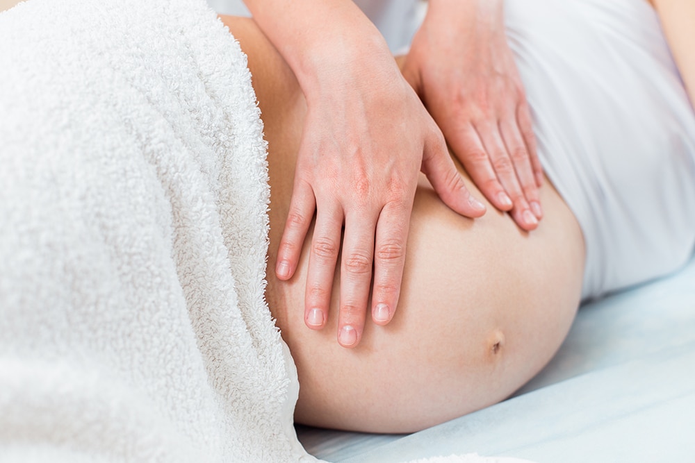 Close-up of a pregnant woman in a white dress sitting on a bed, gently placing her hands on her lower back to relieve discomfort, indicative of the benefits of prenatal holistic massage.