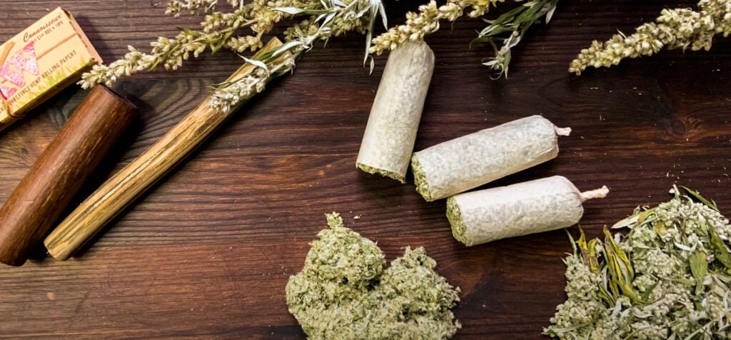 Colocación plana de cogollos de cannabis secos, cigarrillos de hierbas, hierbas esparcidas para moxibustión y un molinillo de madera sobre una superficie rústica de madera.