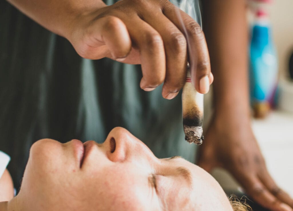 Una mujer acostada con los ojos cerrados mientras otra persona sostiene una vela encendida en su oído, probablemente realizando una terapia de velas para equilibrar el calor.