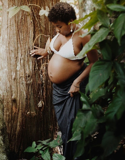 A pregnant woman in a white top and blue skirt stands by a tree in a lush, holistic forest, gently touching her belly and looking thoughtfully down.