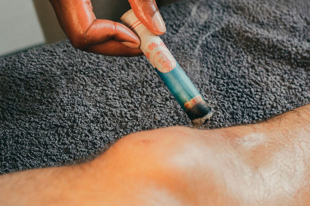 A hand holds a moxibustion bar over a person's knee, suggesting a therapeutic treatment that intertwines with the art of massage. The stick emits smoke and is placed close to the skin. The background features a dark-textured fabric, creating an atmosphere of relaxation and healing.