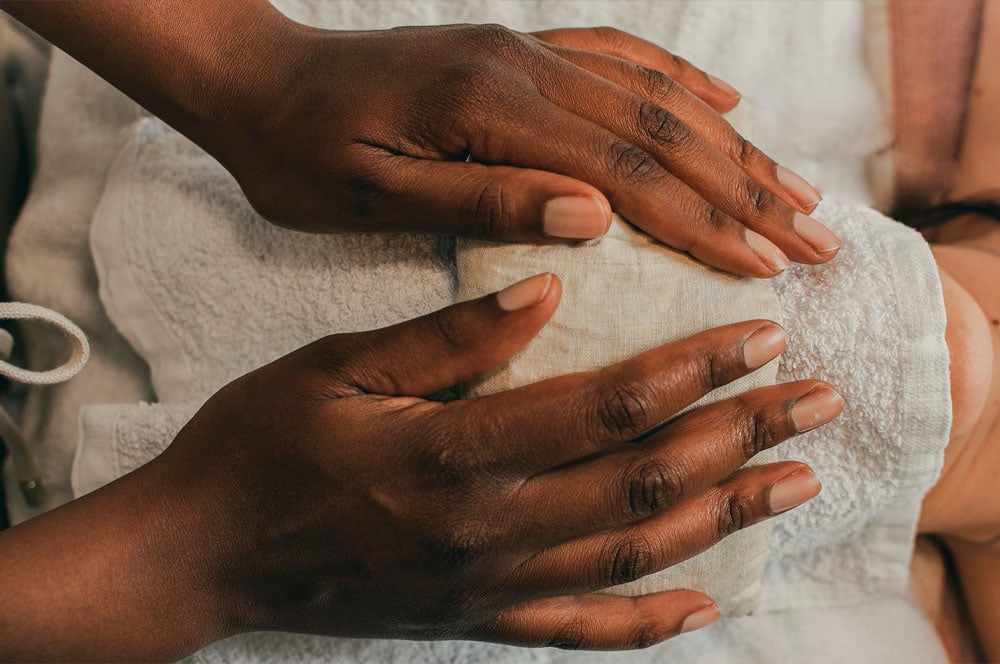 A close-up of a person receiving a spa treatment. A pair of hands are seen gently pressing a warm towel onto the face of another person who is lying down, partially covered with a white towel. The setting oozes calm and relaxation, embodying the art of massage.