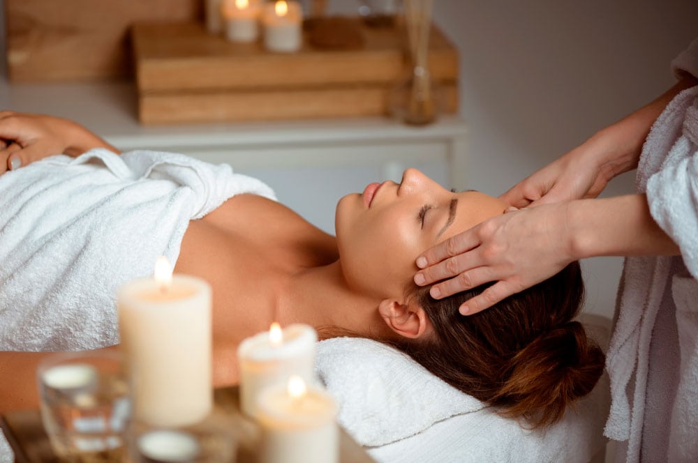 A person lies on a massage table, wrapped in a white towel, while receiving a head massage. In the foreground are several lit candles, which contributes to the relaxing atmosphere. The serene setting enhances the spa atmosphere as if one were experiencing the Art of Massage.