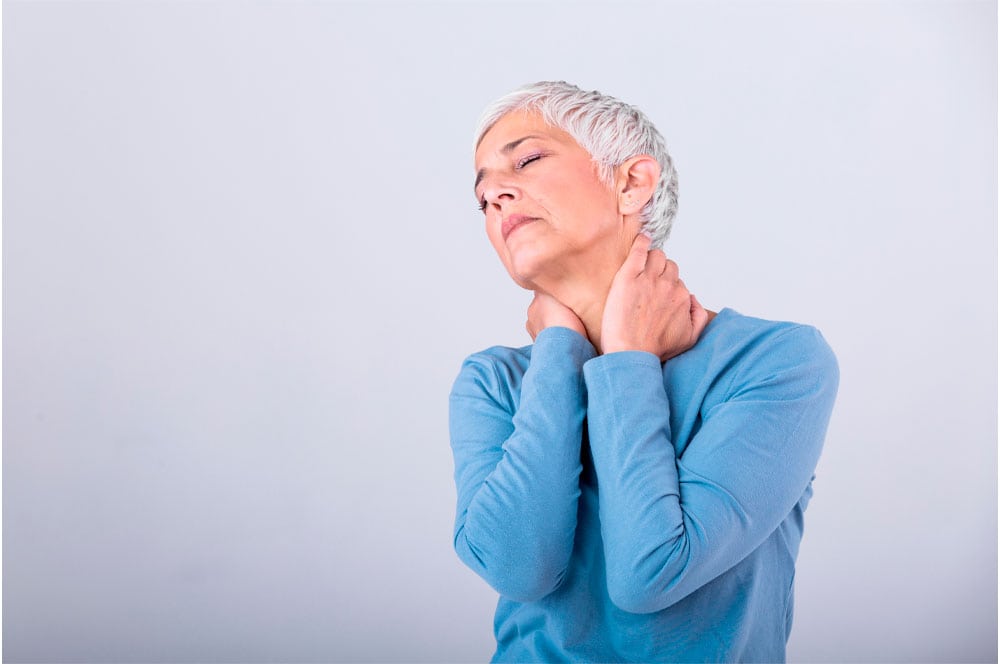 A person with short, silver hair wearing a light blue long-sleeved shirt is tilting their head back slightly while holding their neck with both hands, as if experiencing pain or discomfort, possibly from fibromyalgia. The background is plain and light-colored.