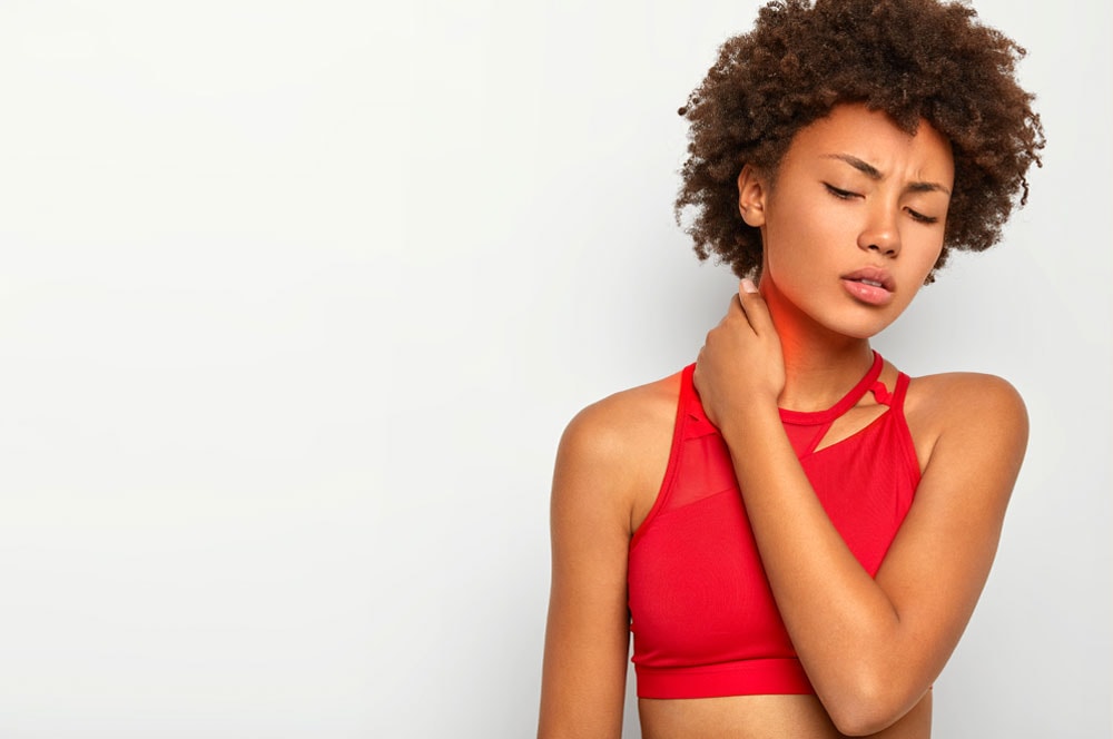 Una persona con pelo rizado y que lleva una camiseta deportiva roja está de pie sobre un fondo liso. Al tocarse el cuello con expresión de dolor, parecen estar desentrañando enredos emocionales, indicando malestar o dolor en la zona del cuello.