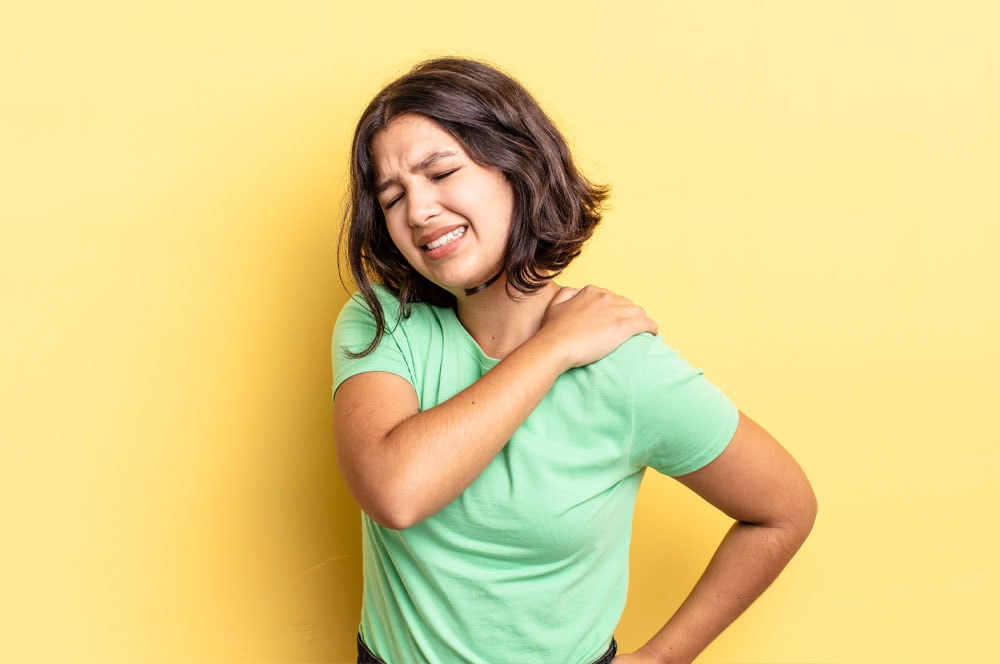 Se ve a una persona con el pelo hasta los hombros, vestida con una camisa verde claro, con expresión de dolor, sujetándose el hombro derecho con la mano izquierda como si sintiera malestar. Están de pie sobre un fondo amarillo, aparentemente desentrañando emociones.