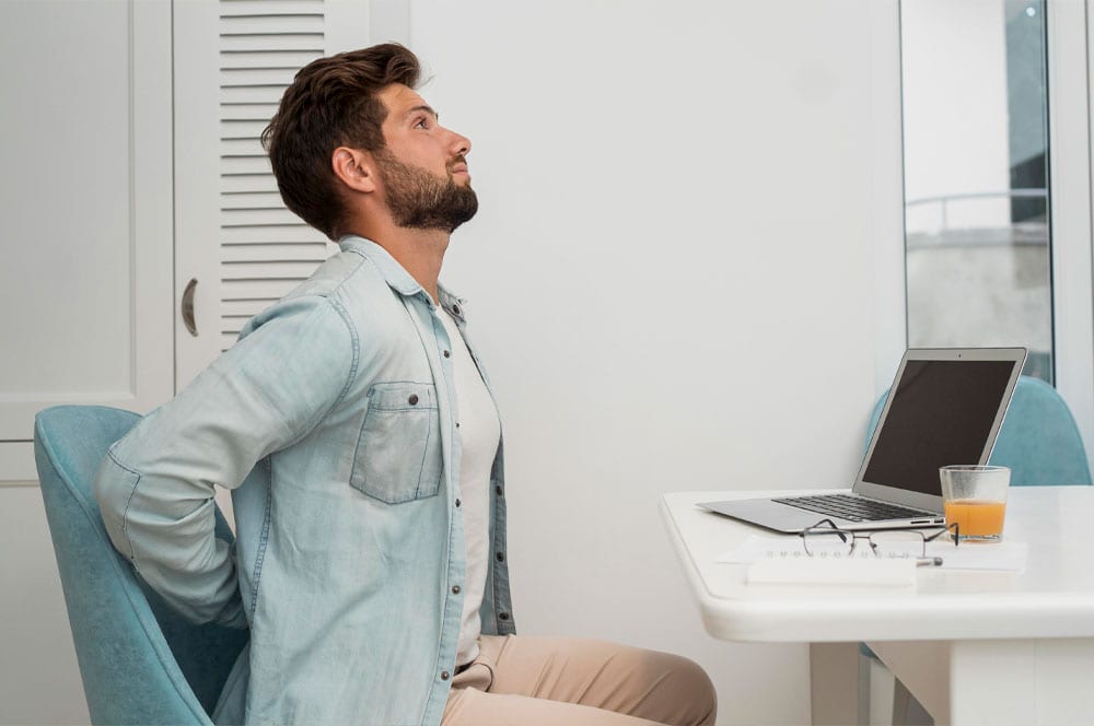 Un hombre con barba y vestimenta informal está sentado en un escritorio con una computadora portátil y un vaso de jugo. Se estira con las manos en la parte baja de la espalda y la cabeza inclinada hacia arriba, desentrañando pensamientos en el espacio interior bien iluminado.