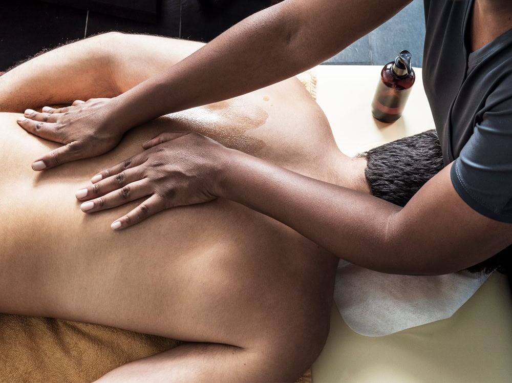 Una persona recibe un masaje de espalda mientras está recostada sobre una camilla de masaje Equilibrio. Las manos del masajista se ven en la espalda de la persona, con aceite de masaje que alinea los chakras visible. Cerca de la mesa hay una botella de este aceite especial.