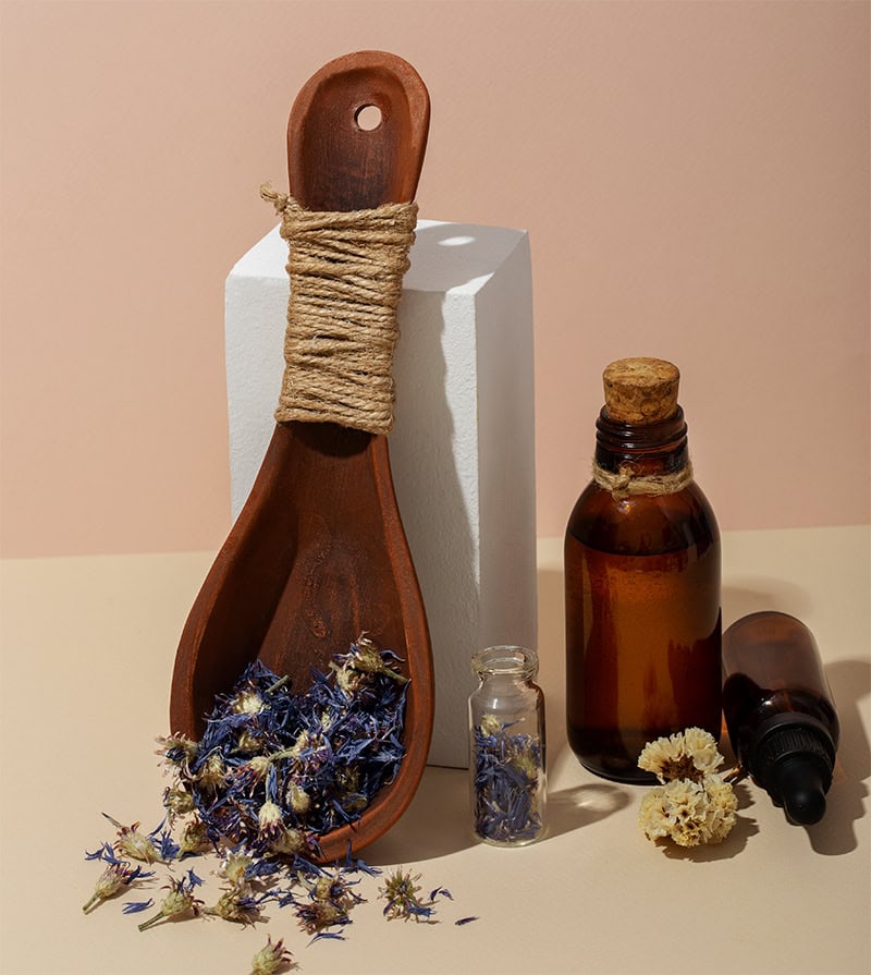 A wooden spoon wrapped with twine and filled with dried flowers, leans against a white pedestal. Nearby are two small glass bottles, one with a dropper, filled with an amber-colored liquid, representing holistic techniques. More dried flowers are scattered on a beige surface.