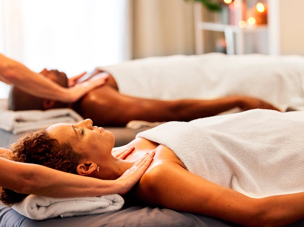 Two people lie side by side on massage tables with white towels covering their bodies. Each person receives a neck massage by a therapist, with a relaxed atmosphere indicated by soft lighting and candles in the background, creating an experience of tranquility and well-being similar to Nirvana.