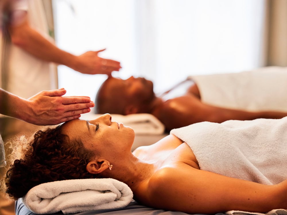 Dos personas relajándose en camillas de masaje, cada una cubierta con una toalla. Un terapeuta, parcialmente visible, realiza un masaje, colocando las manos cerca de la frente de una mujer de pelo rizado en primer plano. La escena es tranquila, bañada por una iluminación suave a través de las ventanas, que ofrece una visión del nirvana.