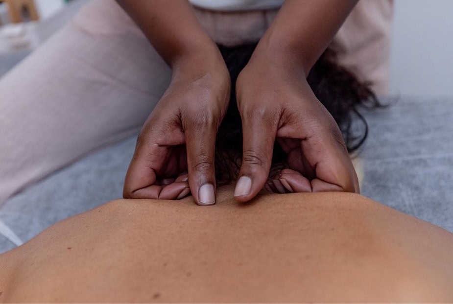 Primer plano de una persona recibiendo un masaje. Se ven dos manos aplicando presión en la parte posterior del cuello de la persona con movimientos firmes y suaves. La persona que recibe el masaje está tumbada boca abajo sobre una camilla de masaje. El fondo está difuminado, centrándose en las manos y la espalda.