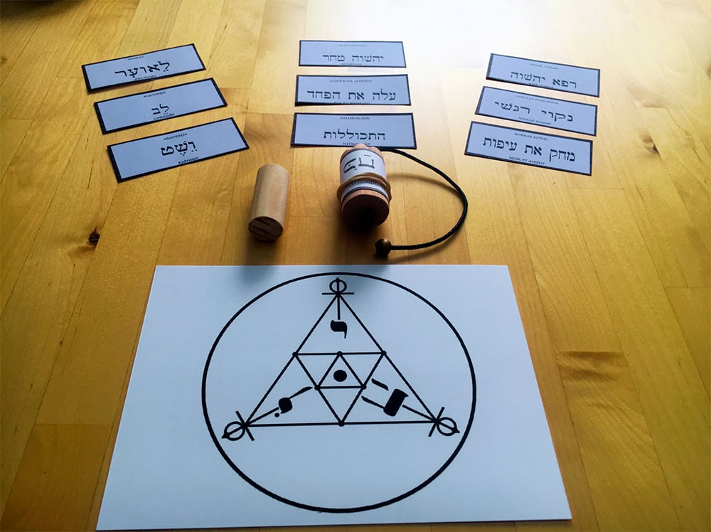 Una mesa de madera con flashcards hebreas, una peonza (dreidel), un pequeño recipiente de madera y una hoja de papel con un diseño geométrico que incorpora letras hebreas. Un péndulo hebreo cuelga cerca, lo que sugiere que la escena está relacionada con el estudio cultural o religioso judío.