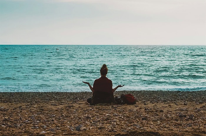 Una persona se sienta con las piernas cruzadas en una playa rocosa frente al océano, practicando una postura meditativa con las manos abiertas y apuntando hacia afuera. El cielo ligeramente nublado contribuye al ambiente sereno, mientras las olas se adentran suavemente. Junto a ellos hay una mochila roja, que se asemeja a un borrador automático de preocupaciones.