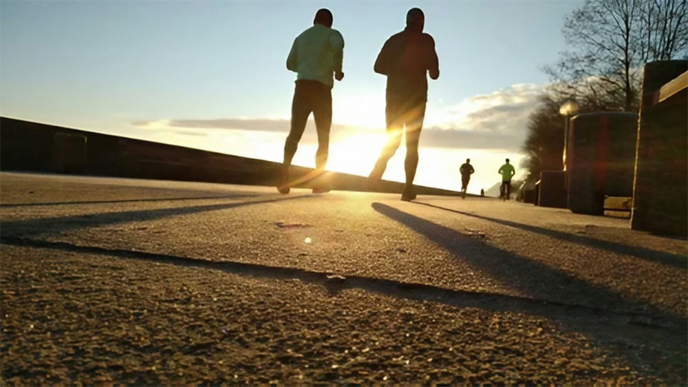 Two people jog along a sunlit trail, casting elongated shadows. The sun is low in the sky, which creates a silhouette effect. Two other corridors can be seen in the distance and trees line the path.