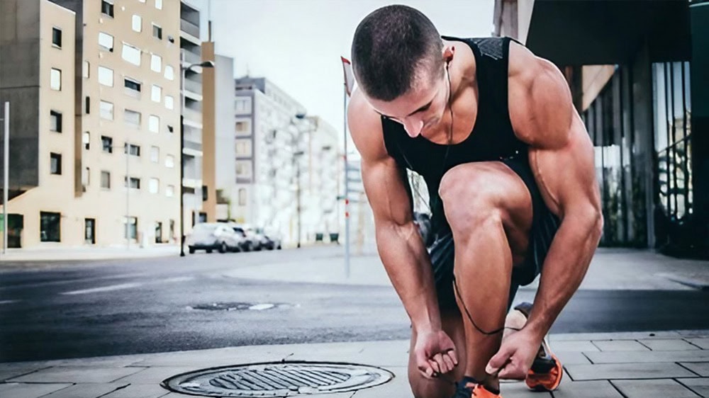 A person in sportswear kneels on an urban sidewalk while tying his sneakers. He wears a tank top and headphones, with buildings and a street in the background.