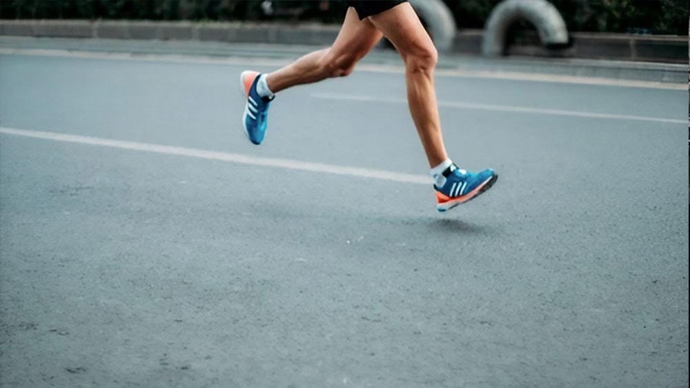 Una persona corre por una carretera vacía con zapatillas azules y naranjas con rayas blancas. Se ven las piernas del corredor, lo que muestra movimiento y velocidad. El fondo incluye una barrera de carretera y vegetación.